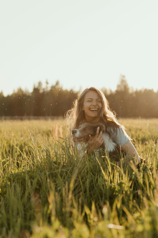 A woman laughing joyfully while embracing a dog in a sunlit field with tall grass.